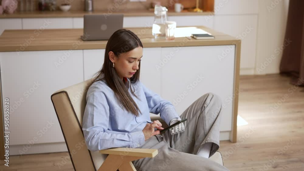 Young disabled woman is holding smartphone and sitting in chair in apartment room spbas. 4k Close view of beautiful american female holds gadget with bionic hand and looks at display, surfs web and Young disabled woman is holding smartphone and sitting in chair in apartment room spbas. 4k Close view of beautiful american female holds gadget with bionic hand and looks at display, surfs web and