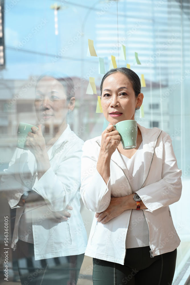 Elegant senior female business leader holding coffee cup and standing near large window at office with city buildings in background Elegant senior female business leader holding coffee cup and standing near large window at office with city buildings in background