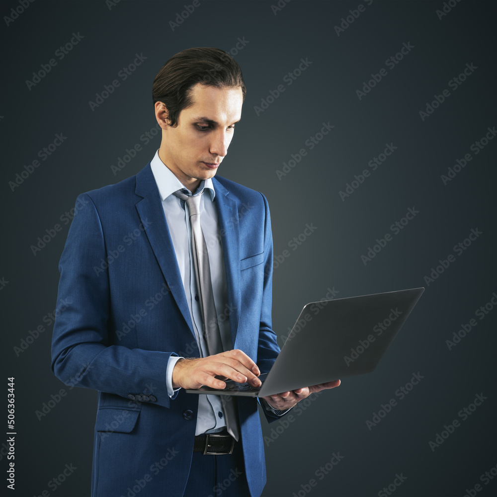 Young businessman in blue suit working with modern laptop, isolated on dark grey wall background, close up Young businessman in blue suit working with modern laptop, isolated on dark grey wall background, close up
