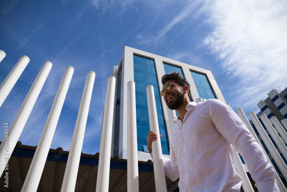 handsome young man with beard, sculpted body and sunglasses is clinging to a white picket fence. The photo is taken from below and you can see the blue sky in the background. handsome young man with beard, sculpted body and sunglasses is clinging to a white picket fence. The photo is taken from below and you can see the blue sky in the background.