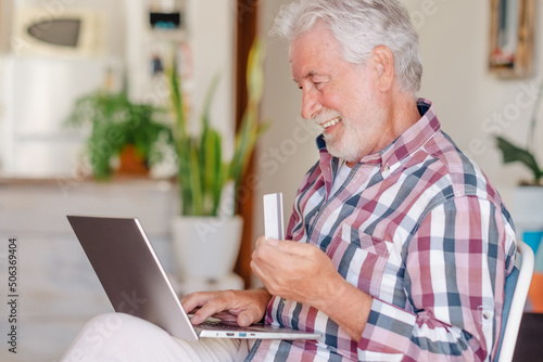 Elderly man browsing by laptop enjoying shopping online. Joyful and smiling beautiful senior grandfather indoors being in great mood using credit card to purchase