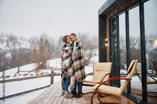 Full length of man and woman wrapped in blanket standing near chairs outside wooden house with panoramic windows. Happy couple hugging outdoors under winter snow. © anatoliy_gleb
