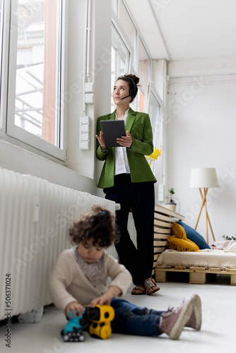 Young businesswoman standing at window working, whilde daughgter is playing on floor photo