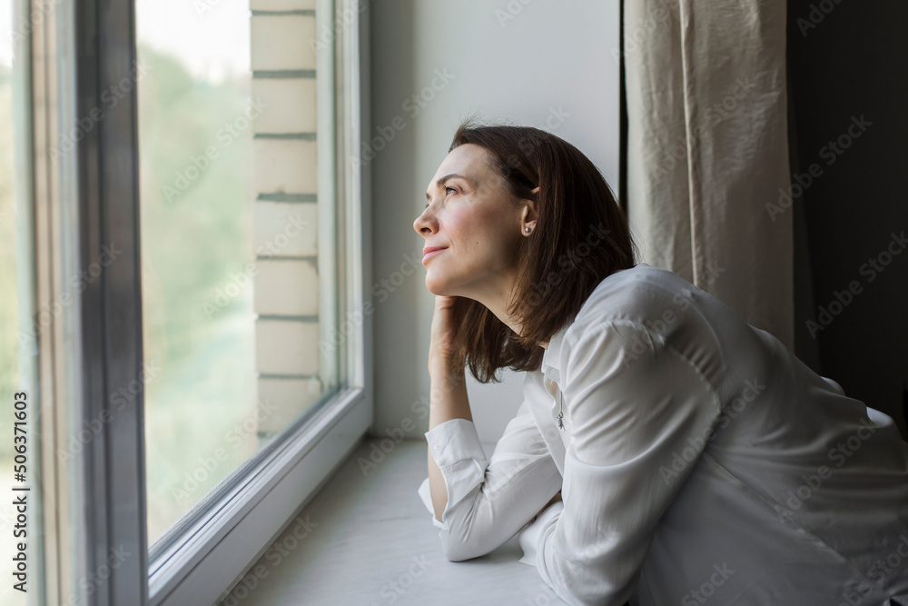 Smiling mature woman looking through window at home Smiling mature woman looking through window at home