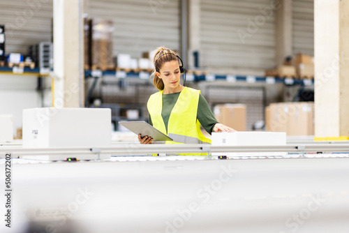 Young worker with tablet PC examining box on conveyor belt in warehouse photo