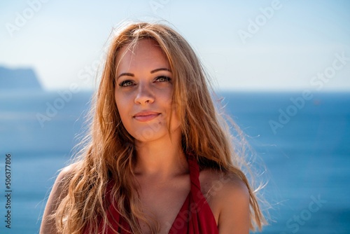 Smiling young woman in a red dress looks at the camera. A beautiful tanned girl enjoys her summer holidays at the sea. Portrait of a stylish carefree woman laughing at the ocean.