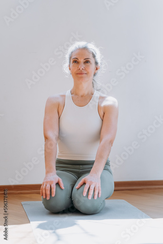 Mature woman practicing yoga asana in room at home. Senior lady sitting at the mat and smiling calmly. Stock photo
