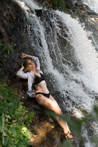 An attractive young woman in a black swimsuit is lying near the waterfall
