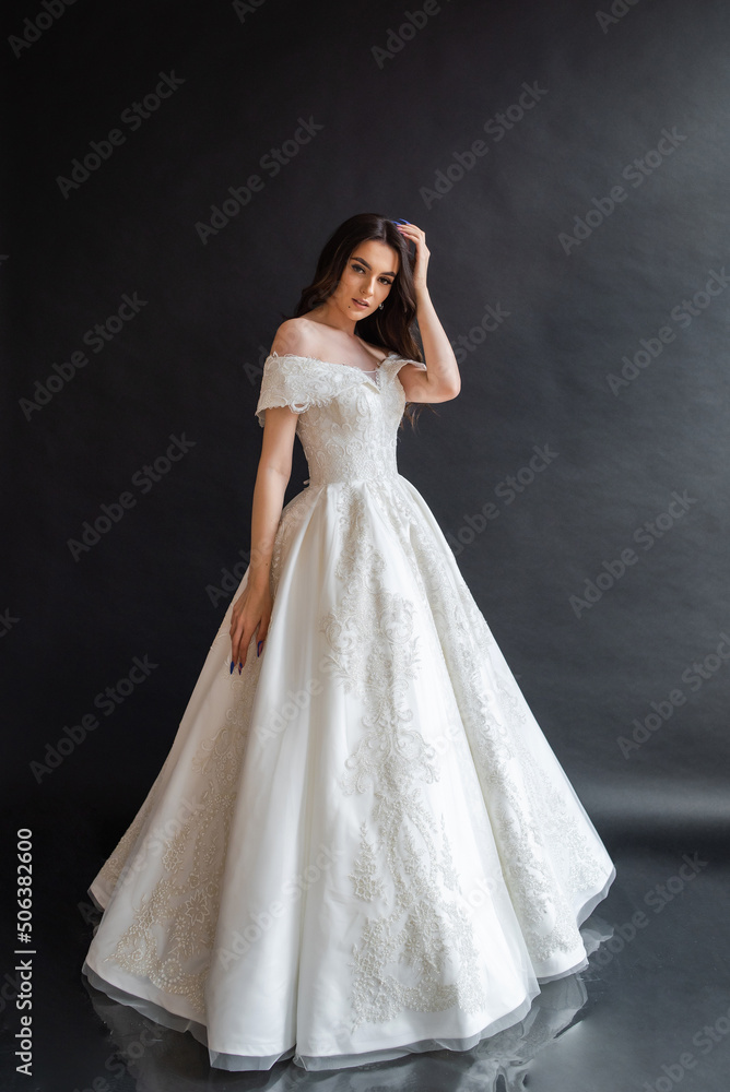 Fototapeta premium Beautiful young sexy model female bride with dark hair in gorgeous glossy long light wedding dress posing in dark studio