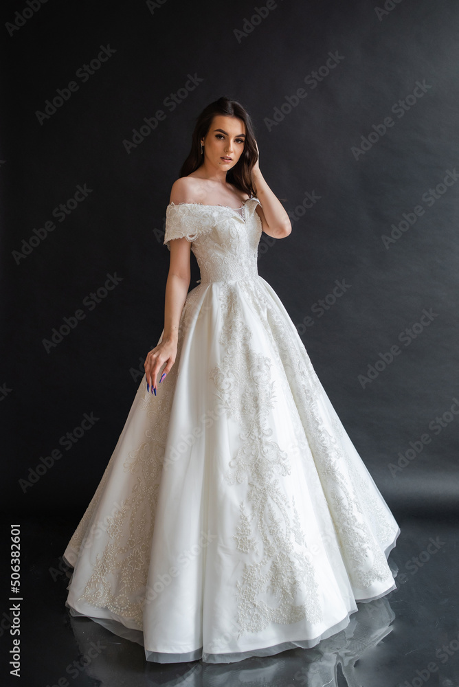 Beautiful young sexy model female bride with dark hair in gorgeous glossy long light wedding dress posing in dark studio