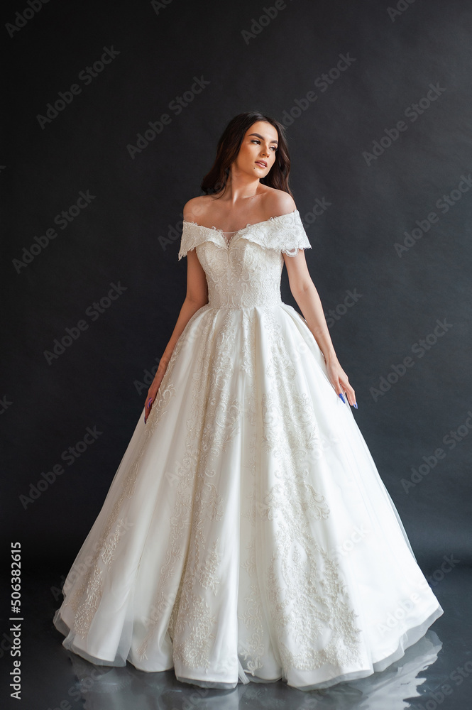 Beautiful young sexy model female bride with dark hair in gorgeous glossy long light wedding dress posing in dark studio