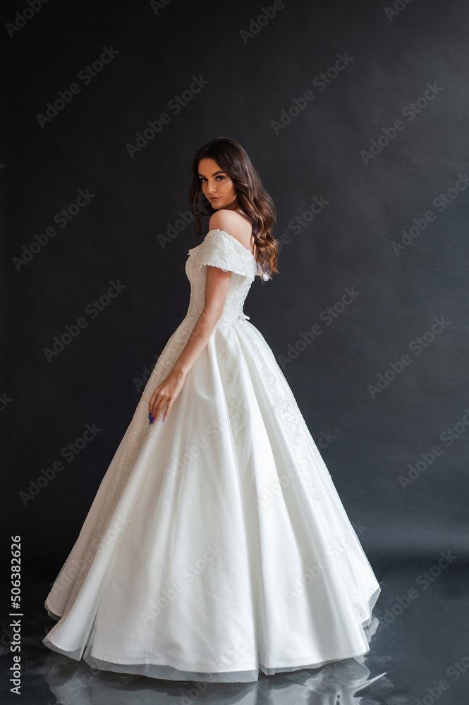 Beautiful young sexy model female bride with dark hair in gorgeous glossy long light wedding dress posing in dark studio