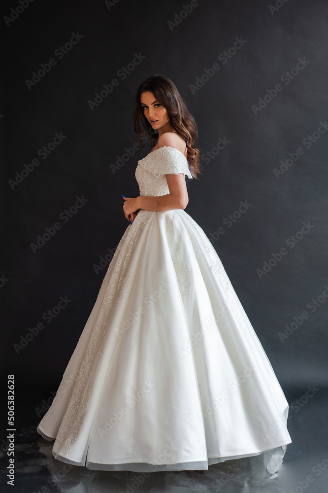 Beautiful young sexy model female bride with dark hair in gorgeous glossy long light wedding dress posing in dark studio Beautiful young sexy model female bride with dark hair in gorgeous glossy long light wedding dress posing in dark studio