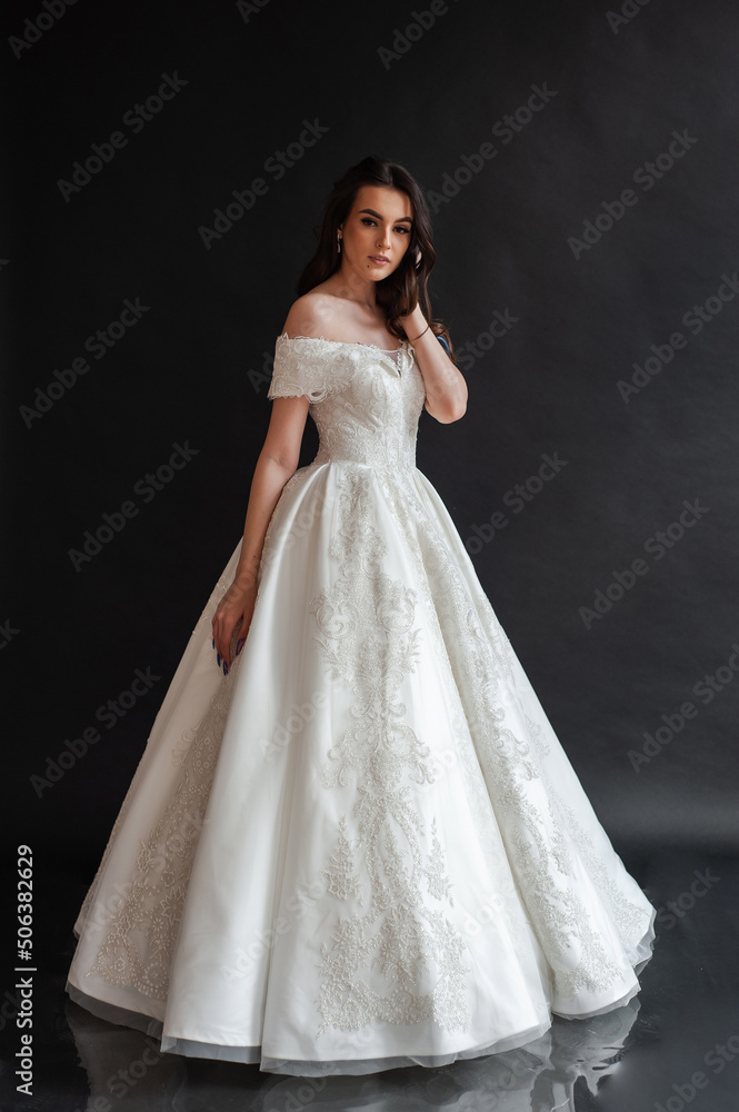 Beautiful young sexy model female bride with dark hair in gorgeous glossy long light wedding dress posing in dark studio Beautiful young sexy model female bride with dark hair in gorgeous glossy long light wedding dress posing in dark studio