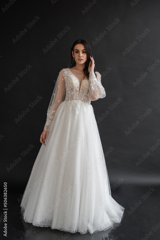 Beautiful young sexy model female bride with dark hair in gorgeous glossy long light wedding dress posing in dark studio