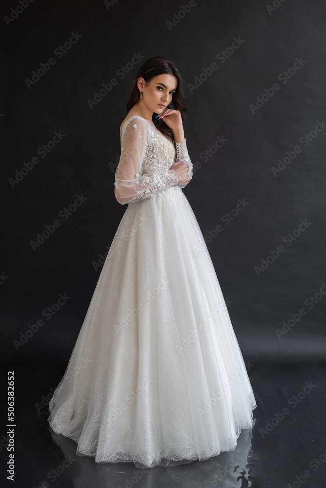 Beautiful young sexy model female bride with dark hair in gorgeous glossy long light wedding dress posing in dark studio
