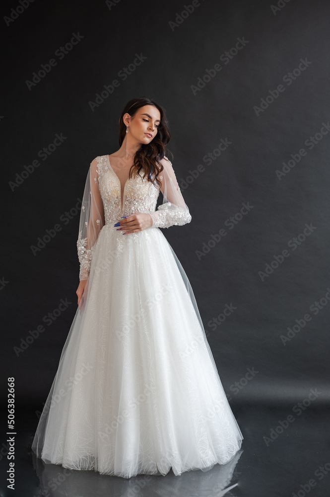 Beautiful young sexy model female bride with dark hair in gorgeous glossy long light wedding dress posing in dark studio Beautiful young sexy model female bride with dark hair in gorgeous glossy long light wedding dress posing in dark studio