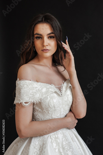 Beautiful young sexy model female bride with dark hair in gorgeous glossy long light wedding dress posing in dark studio