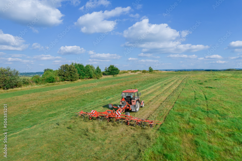 Fototapeta premium A red tractor rakes the mown grass for drying. . Top view of modern machinery on the field.