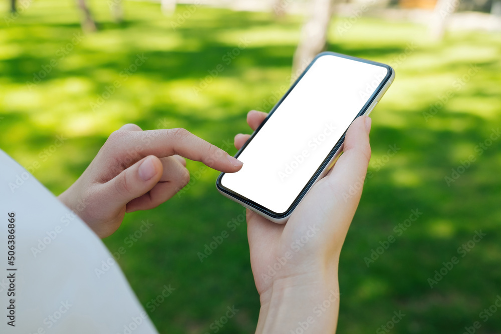 Young woman holding mobile phone with blank white mockup screen on green city park, outdoors. Mobile application tech concept, over shoulder closeup view. Empty screen for social media app.
