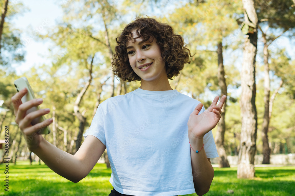 Redhead woman wearing white tee using smartphone video call online. Holding phone looking at screen waving hand video calling distance friend online in mobile chat app using smartphone video chat app. Redhead woman wearing white tee using smartphone video call online. Holding phone looking at screen waving hand video calling distance friend online in mobile chat app using smartphone video chat app.
