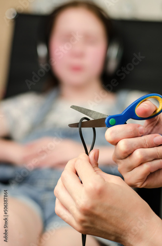 In the foreground, female hands are cutting the headphone cord with scissors, in the background is a girl in headphones. Scissors cut the wire from the headphones