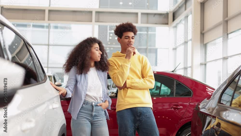 Young couple is choosing new car and talking while standing in bright showroom spbi. Beautiful caucasian woman, man examine transport and talk with smiles, make decision and stand in light interior Young couple is choosing new car and talking while standing in bright showroom spbi. Beautiful caucasian woman, man examine transport and talk with smiles, make decision and stand in light interior