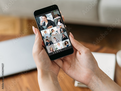 Video chat. Group telecommuting. Distance cooperation. Female hands using phone discussing project online with colleagues on screen.