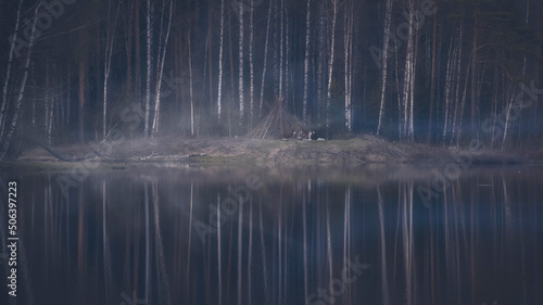 a group of people, friends or tourists on the shore of a foggy mystical lake in the evening near a hut