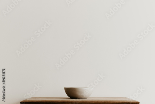 A beige ceramic bowl on a brown wooden board in front of a white wall. Hand made bowl with copy space. Minimal concept, simple design. 