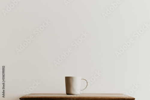 A beige ceramic mug on a brown wooden board in front of a white wall. Hand made coffee cup with copy space. Minimal concept, simple design. 