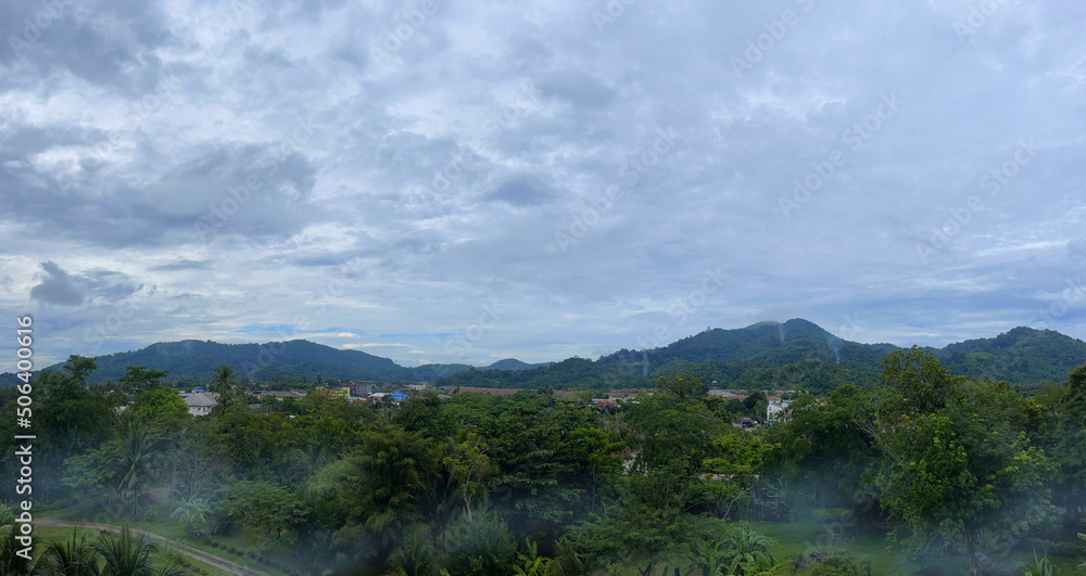 Top view of a green valley, small town and hills on the horizon. Fog creeps across the ground. Wisps of smoke from flattering fires rise in the mountains. Cloudy sky covered with dense clouds. Rainy