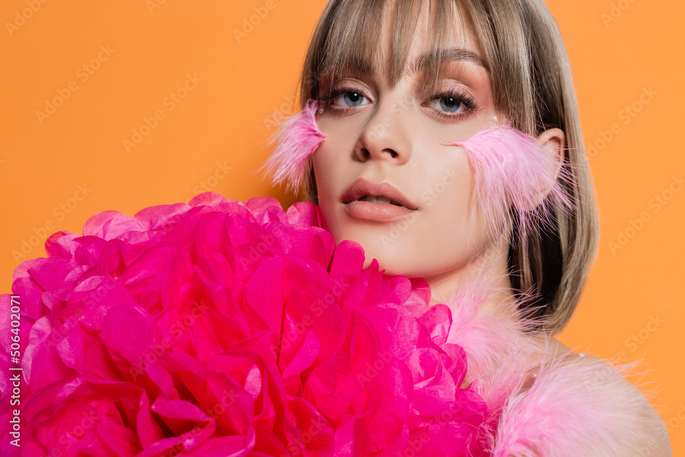 pretty young woman with decorative beads in makeup and feathers on cheeks near pink flower isolated on orange pretty young woman with decorative beads in makeup and feathers on cheeks near pink flower isolated on orange