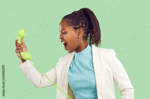 Angry woman annoyed by call and angrily screaming arguing during phone conversation. Young dark skinned woman shouting on colored wired handset from landline retro telephone on light green background.