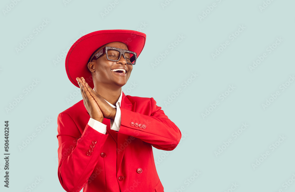 Funny African man in trendy outfit dancing in studio. Happy young Tanzanian guy wearing funky red suit, hat and disco party glasses dancing isolated on blue copy space background. Fashion concept Funny African man in trendy outfit dancing in studio. Happy young Tanzanian guy wearing funky red suit, hat and disco party glasses dancing isolated on blue copy space background. Fashion concept