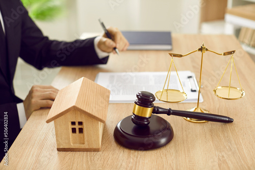 Attorney sitting at desk with scales of justice, gavel and small wooden toy house, working with documents, signing contract agreements. Real estate law, foreclosed property, lawyer services concept photo