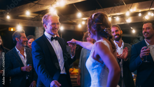 Beautiful Bride in White Dress and Groom in Stylish Black Suit Celebrate Wedding at an Evening Reception Party. Newlyweds Dancing at a Venue with Best Multiethnic Diverse Friends.