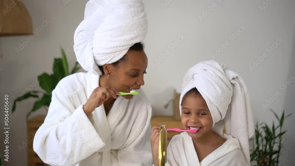 Close view of young mother, cute daughter are brushing teeth and standing in home bathroom spbd. American african woman, girl brush teeth and look with smiles, have fun and stand by sink in light Close view of young mother, cute daughter are brushing teeth and standing in home bathroom spbd. American african woman, girl brush teeth and look with smiles, have fun and stand by sink in light