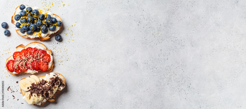 Banner of fruit vegetarian toasts for breakfast on gray background, copy space, top view. Bread slices with ricotta, berries, banana, chocolate and seeds. Banner of fruit vegetarian toasts for breakfast on gray background, copy space, top view. Bread slices with ricotta, berries, banana, chocolate and seeds.