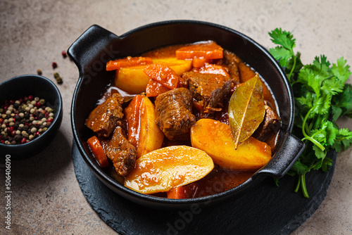 Beef stew with potatoes, carrots in tomato sauce in black pot, dark background.