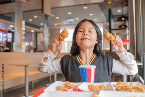 Close up portrait of a satisfied pretty  little asian girl eating fried chicken and french fries  sparkling water in the restaurant. Unhealthy food concept  close up