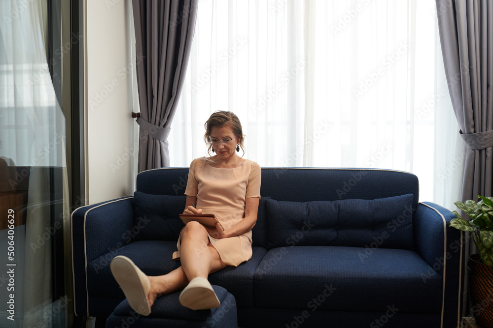 Concentrated middle-aged businesswoman sitting on sofa and proofreading contract on tablet computer Concentrated middle-aged businesswoman sitting on sofa and proofreading contract on tablet computer
