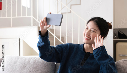 Smiling young asian woman using mobile phone while sitting on a couch at home with laptop computer