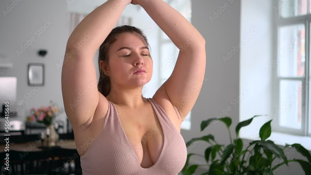 Young overweight woman is practicing yoga and meditating while sitting on floor at home spbd. Close view of oversized Caucasian female does asana with eyes closed and joins hands in mudra, meditates Young overweight woman is practicing yoga and meditating while sitting on floor at home spbd. Close view of oversized Caucasian female does asana with eyes closed and joins hands in mudra, meditates