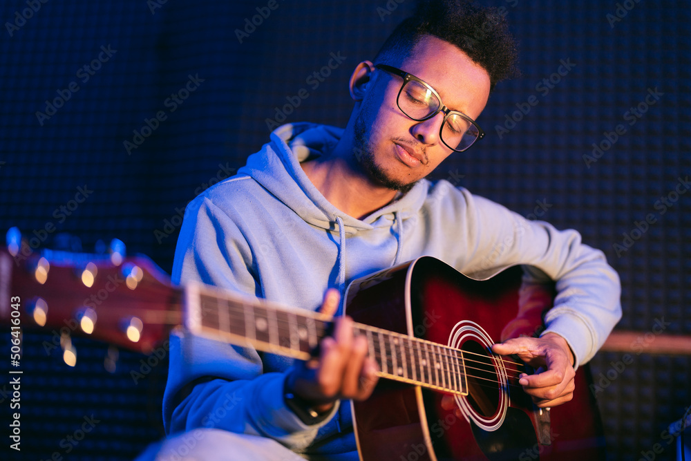 Obraz premium Young afro musician wearing glasses playing acoustic guitar, on dark background