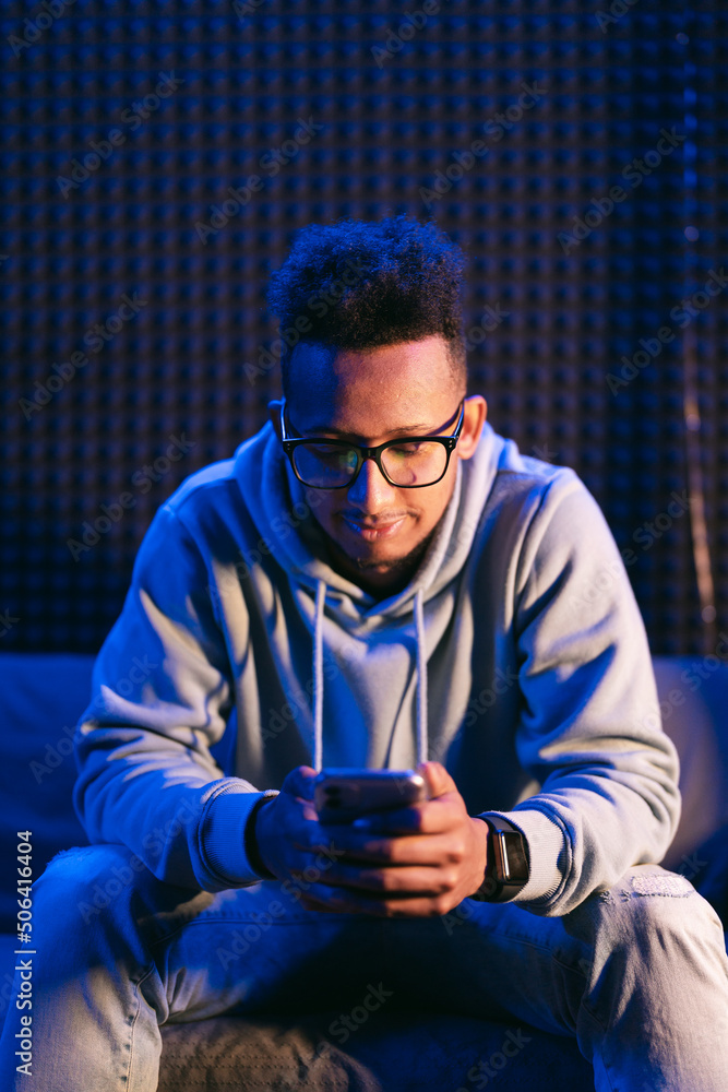 Close-up portrait of an attractive curly guy sitting on sofa using smartphone browsing web service at modern loft industrial style interior splashed by blue light Close-up portrait of an attractive curly guy sitting on sofa using smartphone browsing web service at modern loft industrial style interior splashed by blue light