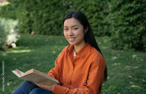 Beautiful smiling Vietnamese student studying, reading book looking at camera sitting in park. Education concept