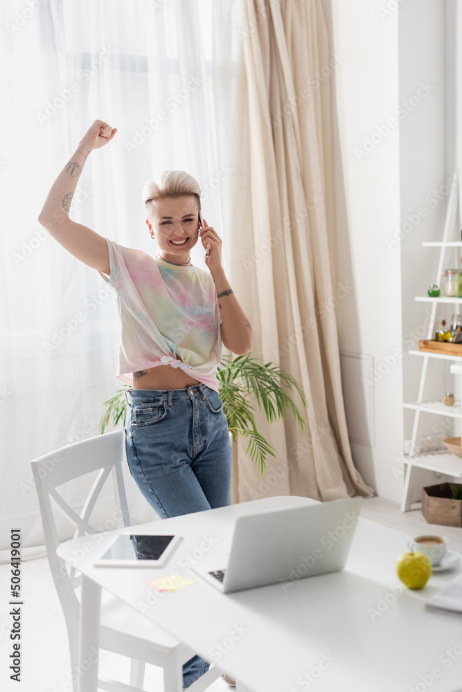 excited woman showing triumph gesture while talking on smartphone near laptop in kitchen excited woman showing triumph gesture while talking on smartphone near laptop in kitchen