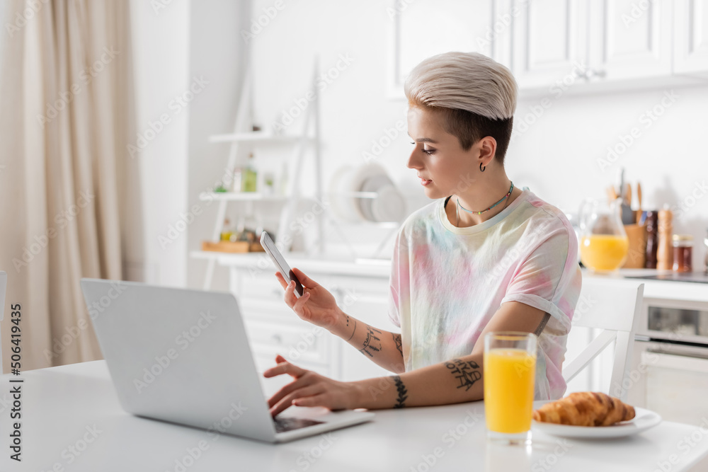 trendy tattooed woman with mobile phone typing on laptop near orange juice and croissant