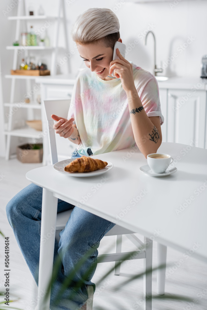 joyful woman talking on smartphone near delicious croissant and coffee cup